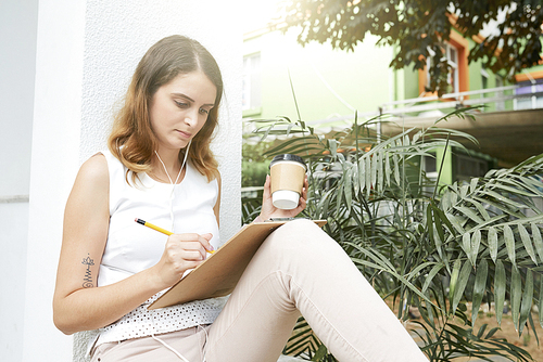 Pretty young woman drinking take-away coffee and taking notes in planner