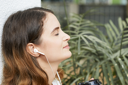 Pretty young woman sitting outdoors and closing her eyes when listening to music in her earphones