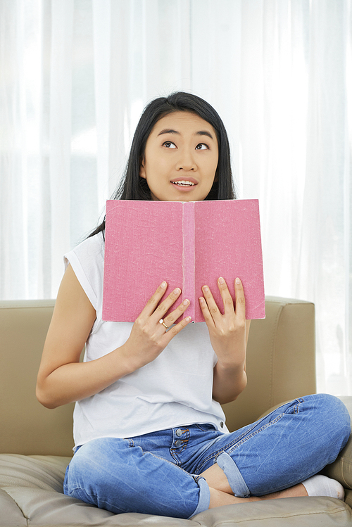 Portrait of positive Asian girl sitting on sofa with opened book and trying to memorize material for school