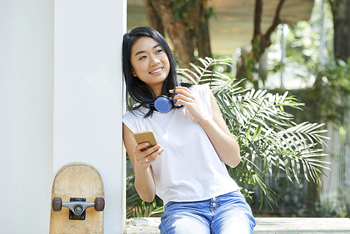 Portrait of pretty Vietnamese teenage girl with smartphone and headphones resting outdoors on summer day