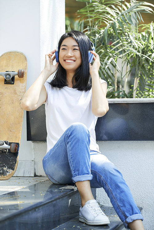 Positive Vietnamese teenage girl sitting on steps and listening to music in her headphones