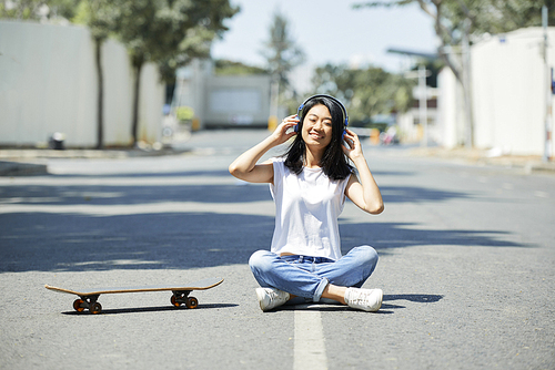 Pretty happy Asian teenage girl sitting on road next to skateboard and listening to music in her headphones