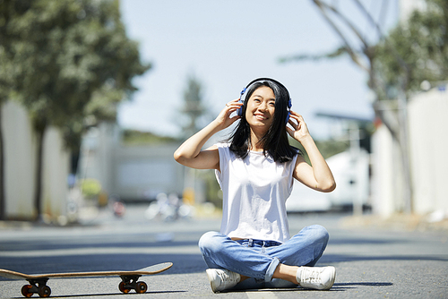 Happy smiling Vietnamese teenage girl wearing headphones sitting on ground and listening to music