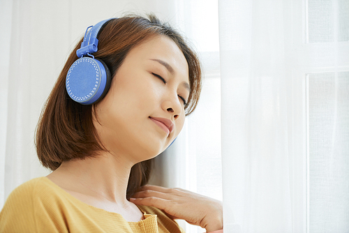 Young Asian woman sitting near window, closing eyes and enjoying calm relaxing music in headphones