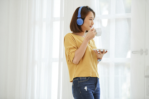 Pretty young Vietnamese woman in big headphones standing near window and drinking cup of tea