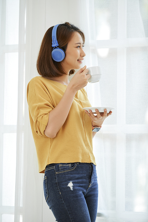 Pretty young Asian woman drinking morning coffee and looking through big window in her room