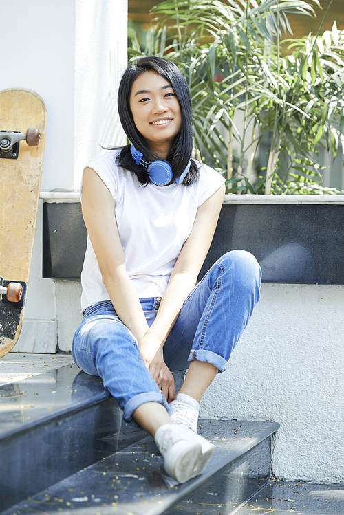 Portrait of pretty smiling Asian teenage girl resting on steps with skateboard next to her