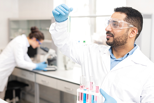 Content experienced young male chemist in lab coat holding rack and analyzing sample in test tube while working in modern laboratory