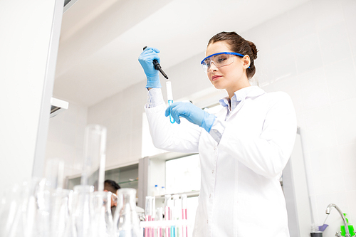 Content experienced young lady chemist in lab coat using dropper and test tube while mixing reagents in laboratory