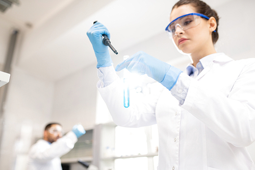 Below view of concentrated young woman in white coat and rubber gloves using dropper while mixing chemicals in test tube