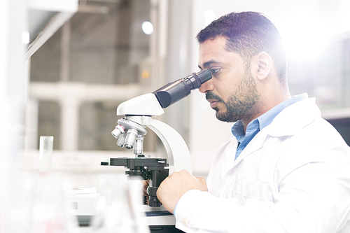 Serious focused handsome young Arabian biologist sitting at desk and using microscope while analyzing sample of modified gene