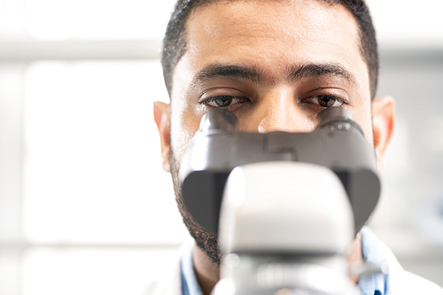 Close-up of focused young Arabian lab biologist looking through microscope to enlarge sample while studying cells in laboratory