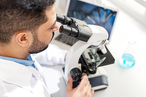 Serious busy young middle-eastern scientist sitting at table with flask and laptop and using microscope while working with samples in laboratory