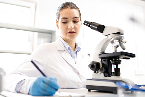 Serious thoughtful young woman in lab coat sitting at table and making records about research in laboratory