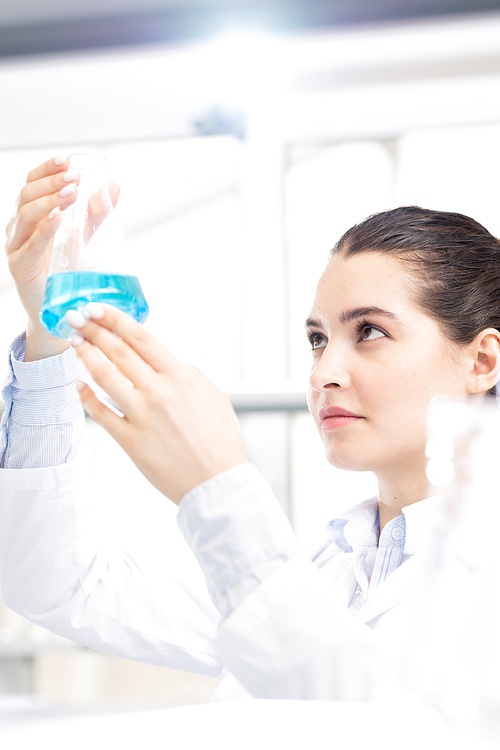 Serious thoughtful young female chemist in lab coat holding flask up while checking chemical liquid in laboratory