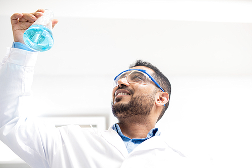 Below view of cheerful confident young middle-eastern chemist with beard satisfied with test solution holding flask with blue liquid substance