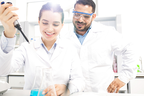 Cheerful optimistic multi-ethnic chemists in white coats performing experiment in laboratory: smiling woman dropping reagent into flask