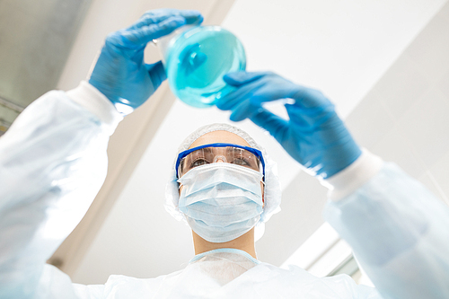 Directly below view of serious concentrated lady chemist in protective mask and goggles holding flask and examining color of liquid in laboratory