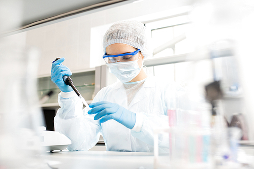 Serious concentrated female microbiologist in sterile clothing and safety goggles sitting at table and dropping reagent in petri dish while doing research in laboratory