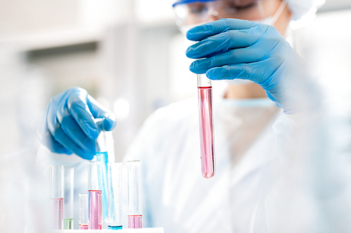 Close-up of busy woman in rubber gloves putting test tubes with colorful liquids into rack while working with chemical substances in laboratory