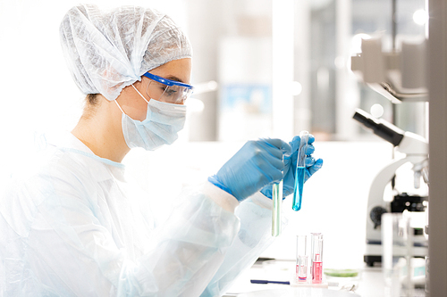 Serious focused young female clinical laboratory researcher in protective coat, gloves and mask sitting at desk and comparing chemical liquids in test tubes