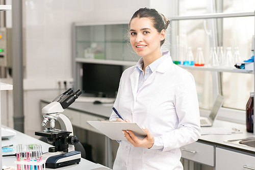 Smiling attractive young lady researcher in lab coat standing in laboratory and making notes about chemicals in clipboard, she 