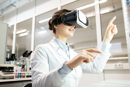 Content inquisitive young female laboratory worker in white coat pointing with fingers while watching video on VR device