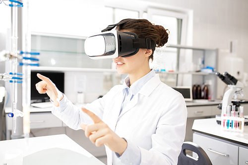 Smiling smart young female scientific researcher gesturing hands while using virtual reality simulator in laboratory