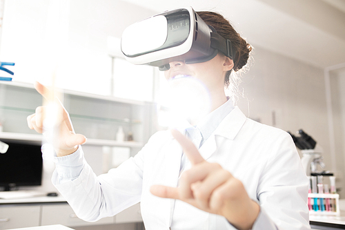 Positive curious modern young female scientist in white coat using invisible monitor while learning about genes with VR simulator