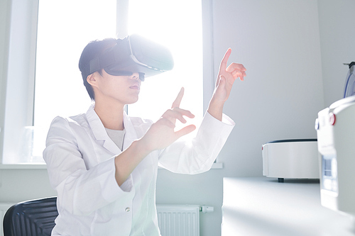 Serious concentrated young Asian innovation scientist in lab coat sitting at table and gesturing hands while using VR simulator for studying cells in 3D