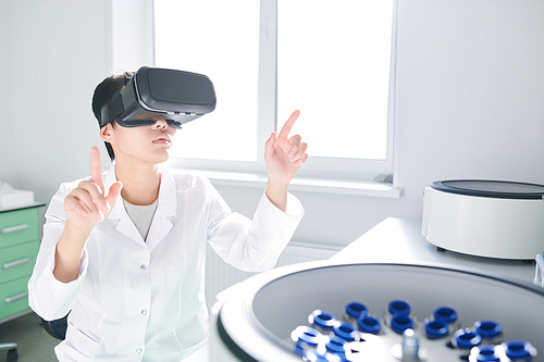 Serious thoughtful young Asian laboratory technician in white coat sitting at table and pointing with fingers while synchronizing VR simulator with centrifuge