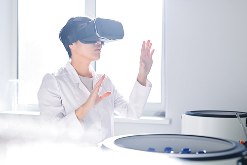 Concentrated young Asian laboratory scientist in white coat gesturing hands and using virtual reality simulator while studying cell and bacteria