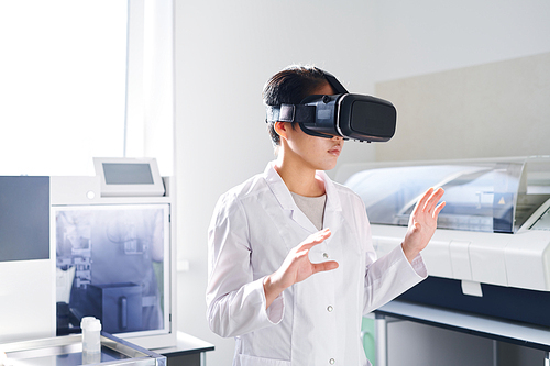 Serious thoughtful young Asian laboratory scientist with short hair standing in modern laboratory and using virtual reality simulator while watching educational video