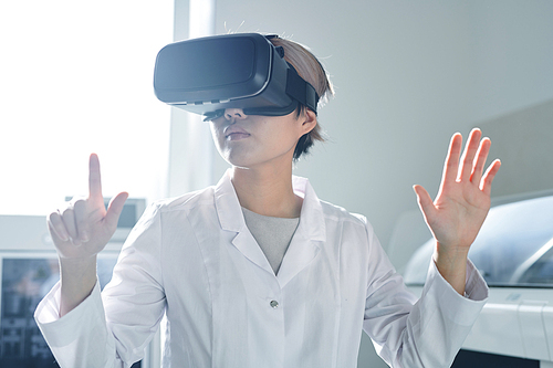 Serious confident young Asian lady scientist in white coat editing research information on invisible board while augmented reality simulator in laboratory