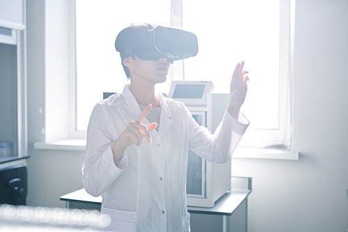 Serious concentrated young Asian laboratory technician in white coat gesturing hands while viewing information in 3D via virtual reality simulator, she doing innovation research