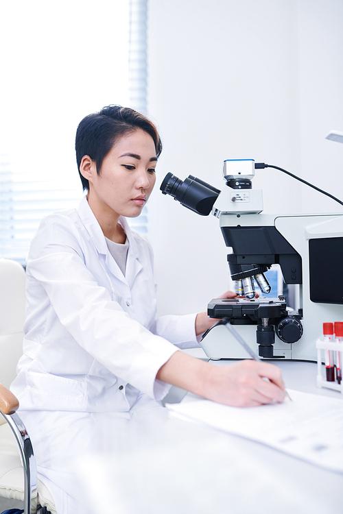 Serious thoughtful young Asian biologist in white coat sitting at table and recording data while studying cells with microscope