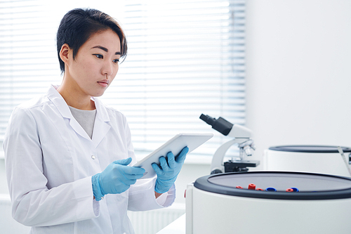 Serious concentrated young Asian modern researcher in lab coat standing at centrifuge and viewing results of test on tablet