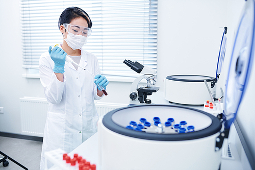 Serious concentrated young Asian laboratory researcher with short hair wearing white coat, mask and safety goggles mixing reagent with blood sample at workplace