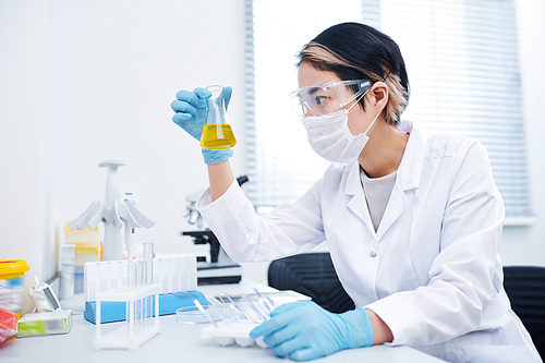 Serious concentrated young Asian female chemist in mask and protective goggles sitting at table and holding flask of medical sample, she working with fluids