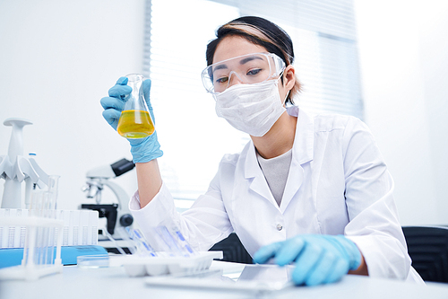 Busy young Asian female lab technician in safety goggles and surgical mask sitting at table and processing specimen while doing medical test