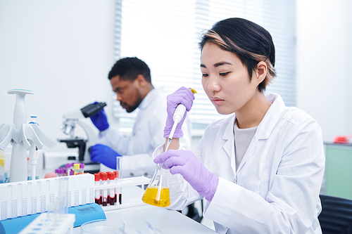 Concentrated young Asian female lab technician with short hair sitting at desk and dropping yellow liquid into flask while testing sample