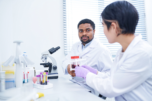 Serious thoughtful multi-ethnic laboratory workers in white coats sitting at table and discussing investigation of samples, Asian lady giving test tube rack to colleague