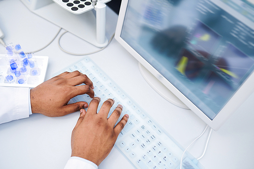 Close-up of unrecognizable black laboratory researcher sitting at table and typing on computer keyboard while using computer program for modelling genes