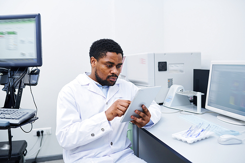 Serious concentrated young black medical scientist with beard sitting at desk and using digital tablet while analyzing data in laboratory