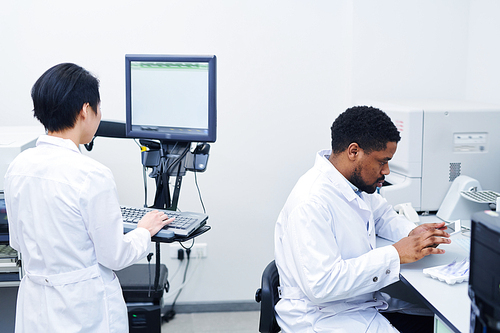 Hospital specialists working on modern equipment in laboratory: serious black scientist using mobile app on tablet while Asian lady working with computer