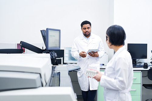 Serious young black doctor in lab coat standing at desk and using tablet while talking to colleague about laboratory research