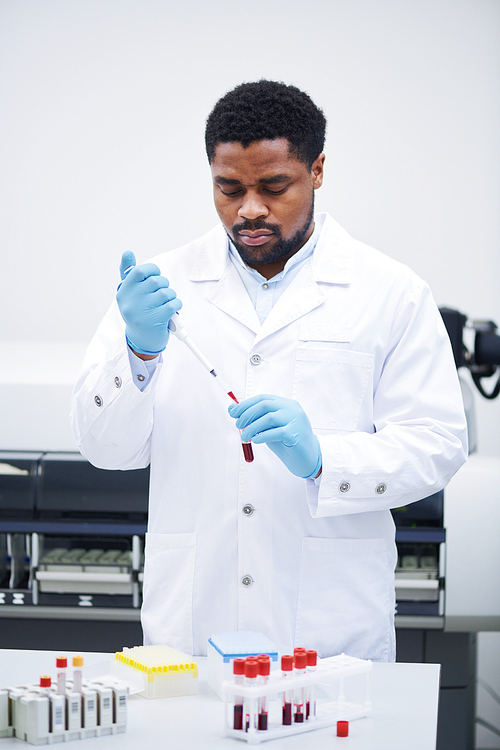 Serious concentrated young African-American medical specialist in white coat standing at table and dropping blood into test tube while analyzing blood sample in laboratory