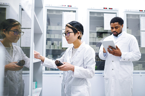 Serious young Asian pharmacist in lab coat and glasses standing at cabinet and sorting medications in storage room