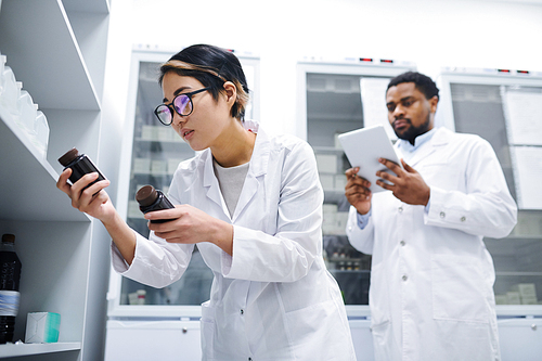 Concentrated young Asian lady doctor in lab coat standing at cabinet and checking medications while reading labels on pill bottles