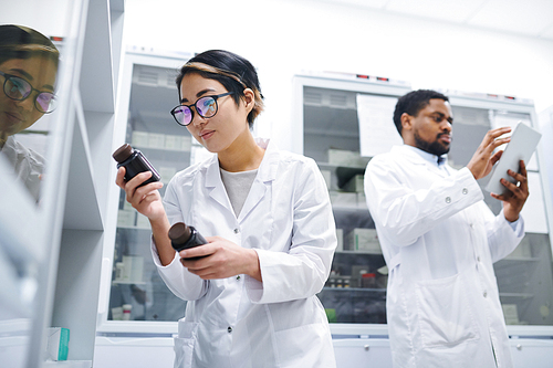 Serious concentrated young Asian female pharmacist with short hair standing at refrigerator with medications and finding necessary pills in medical storage room